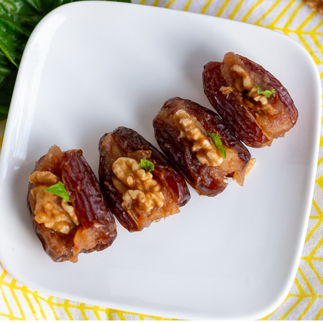 2 Dates stuffed with applesauce, walnuts, and topped with basil sitting on a white rectangular plate with rounded corners. yellow placemat underneath plate.